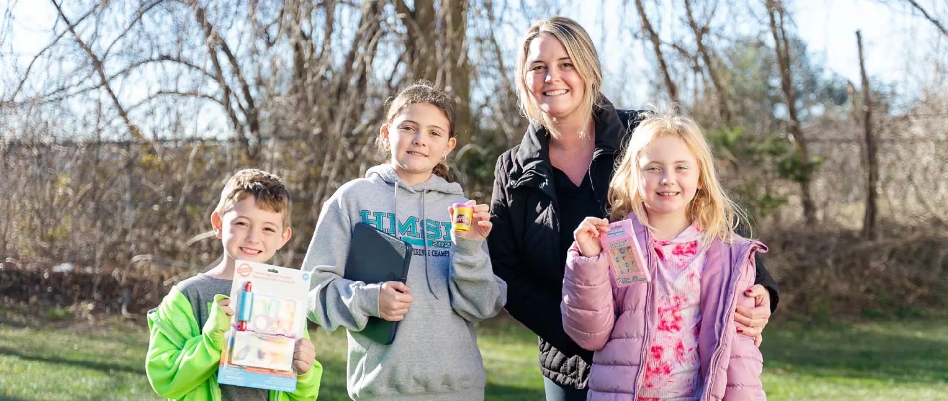 A smiling woman and three children standing outdoors in a yard, holding up various toys and small items from Howell Pediatric Dentistry